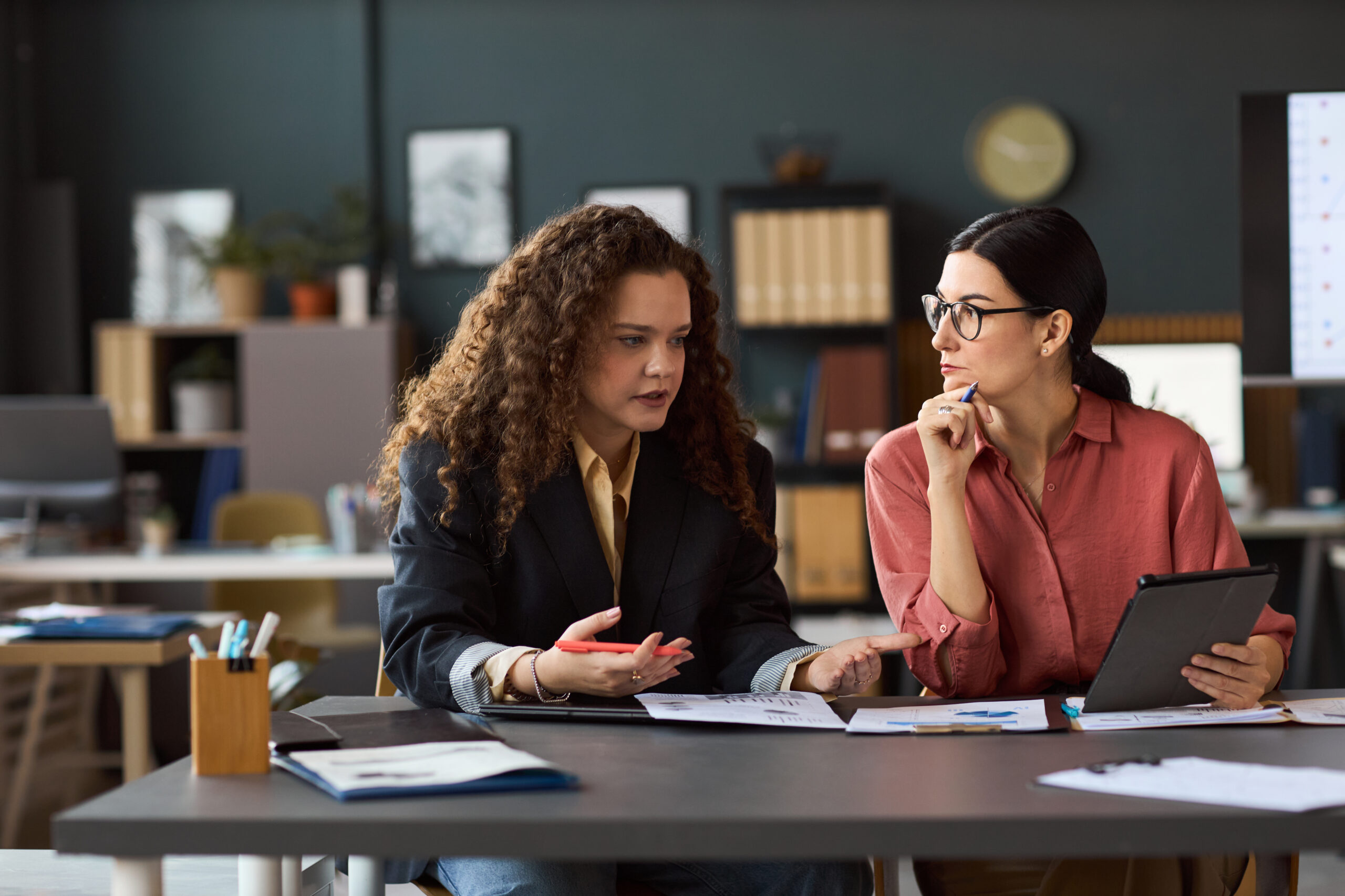 Two young adult women sitting at a desk discussing in a modern office workspace