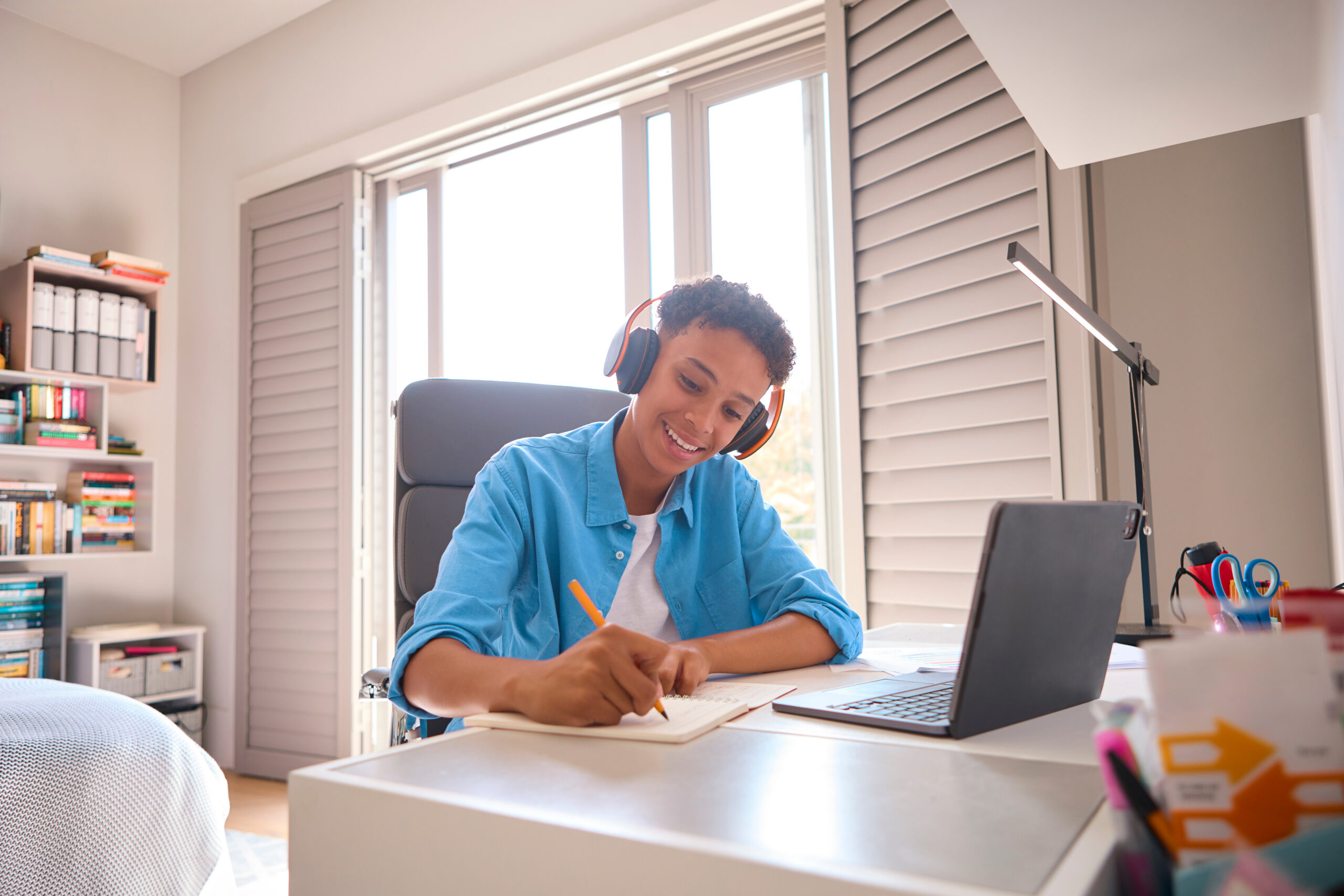 Happy teenage boy writing at desk next to laptop at home.